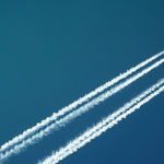 A plane cuts diagonally across a deep blue sky, leaving contrails behind it